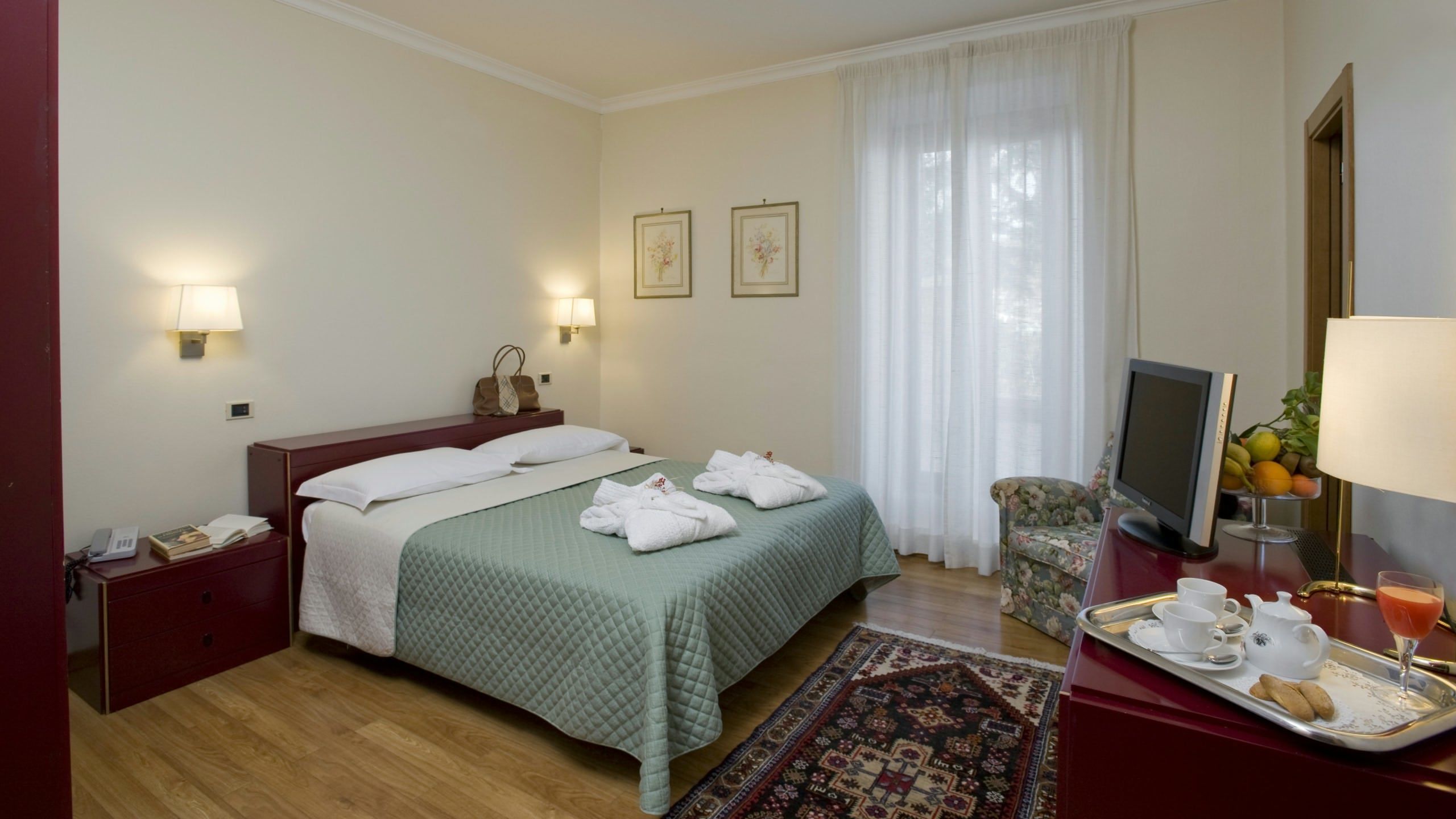 A hotel room with a bed, bedside tables, a desk with a computer, a chair, and a tray with breakfast items.
