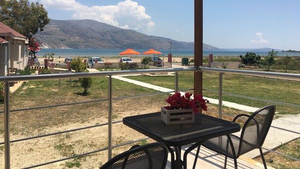 A balcony with a table and chairs, overlooking a beach with umbrellas and a car. - Daphne's Boutique Apartments (Kefalonia)