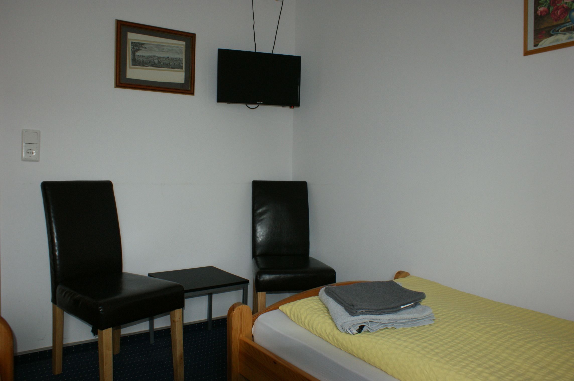 A small room with a bed, two chairs, a desk, a television, and a framed picture on the wall.