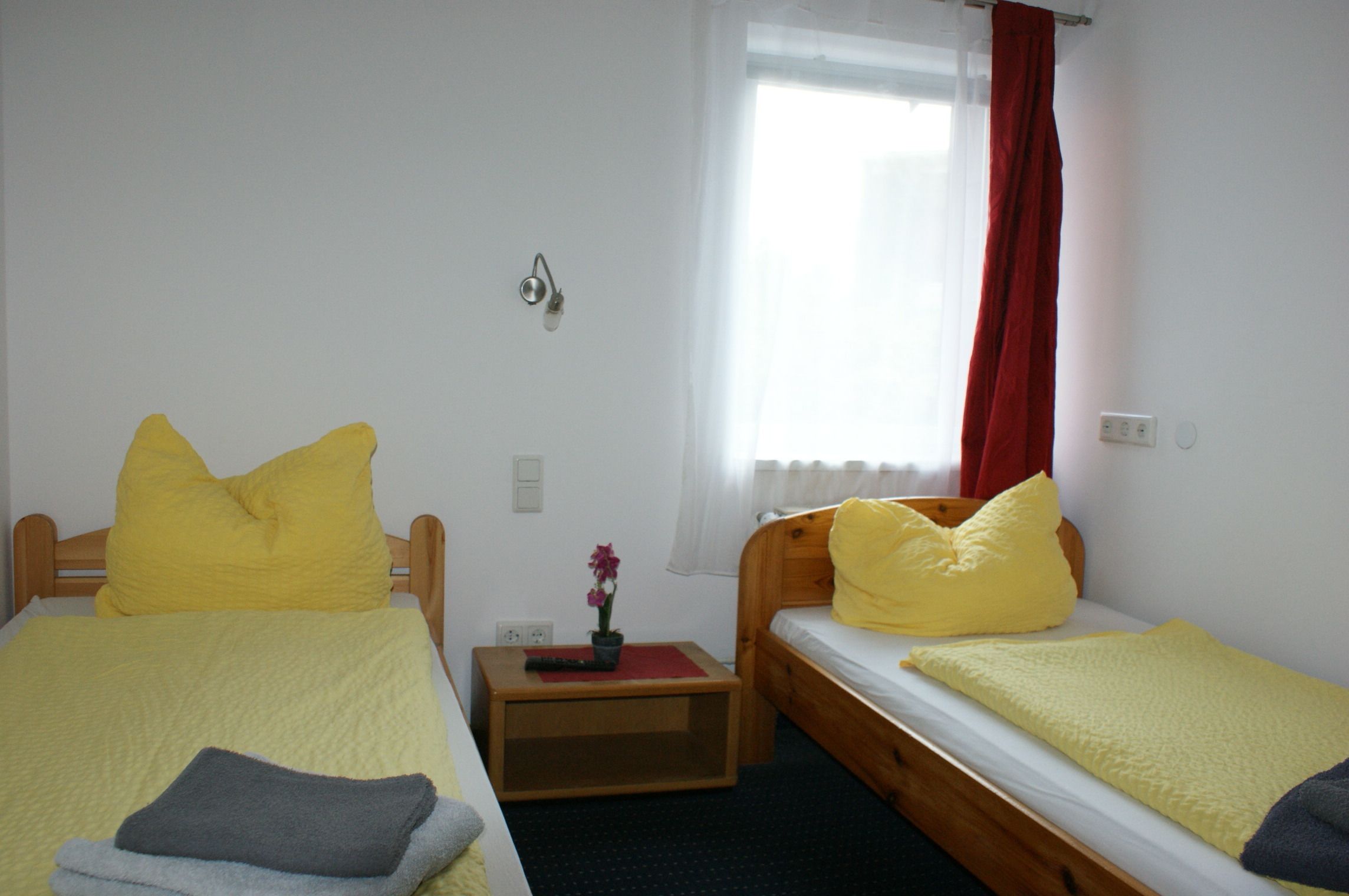 A room with two single beds, each with a yellow bedspread and pillows. There is a small wooden nightstand with a remote control and a potted plant. A window with a red curtain is visible.