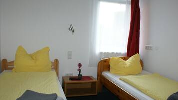 A room with two single beds, each with a yellow bedspread and pillows. There is a small wooden nightstand with a remote control and a potted plant. A window with a red curtain is visible.