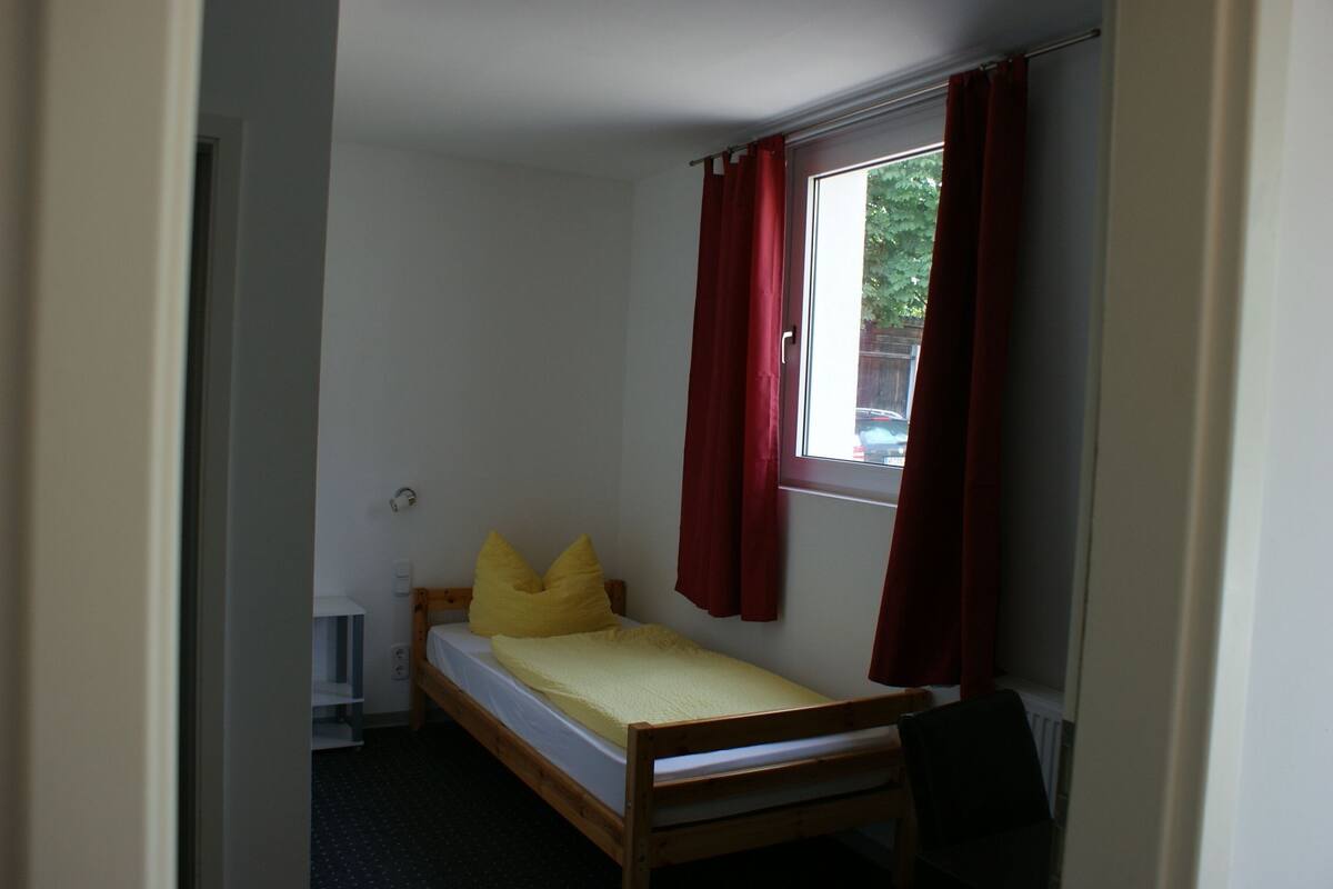 A small, single-bed room with a window, a radiator, and a chair.