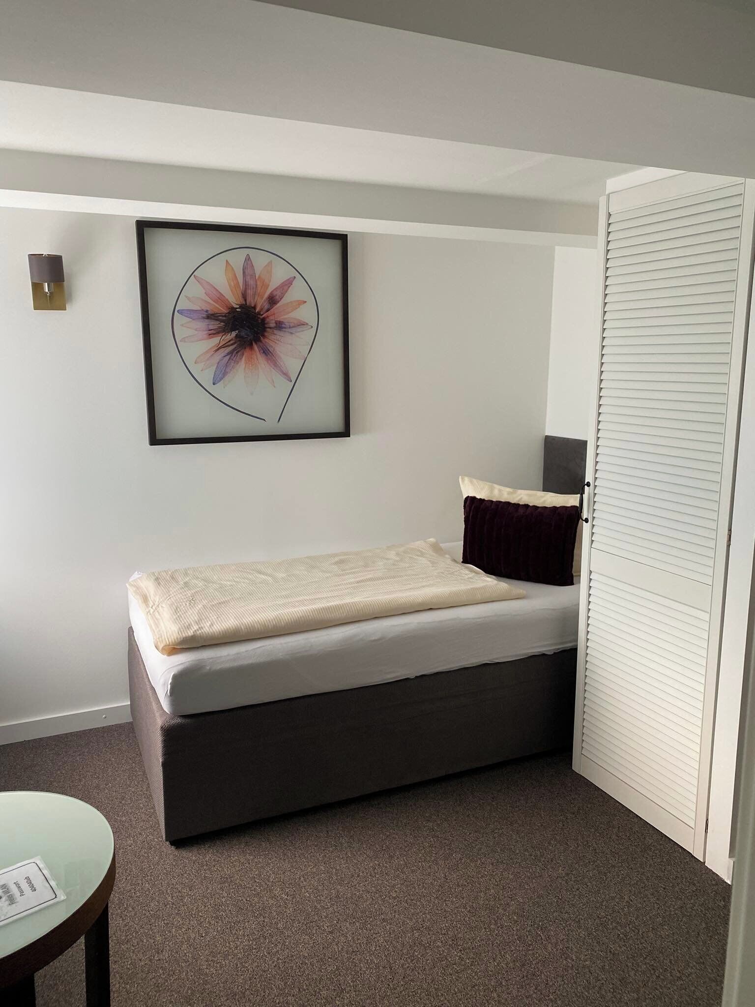 A small, single-bed room with a grey mattress, a dark brown bench, a framed floral artwork on the wall, and a closed white door.