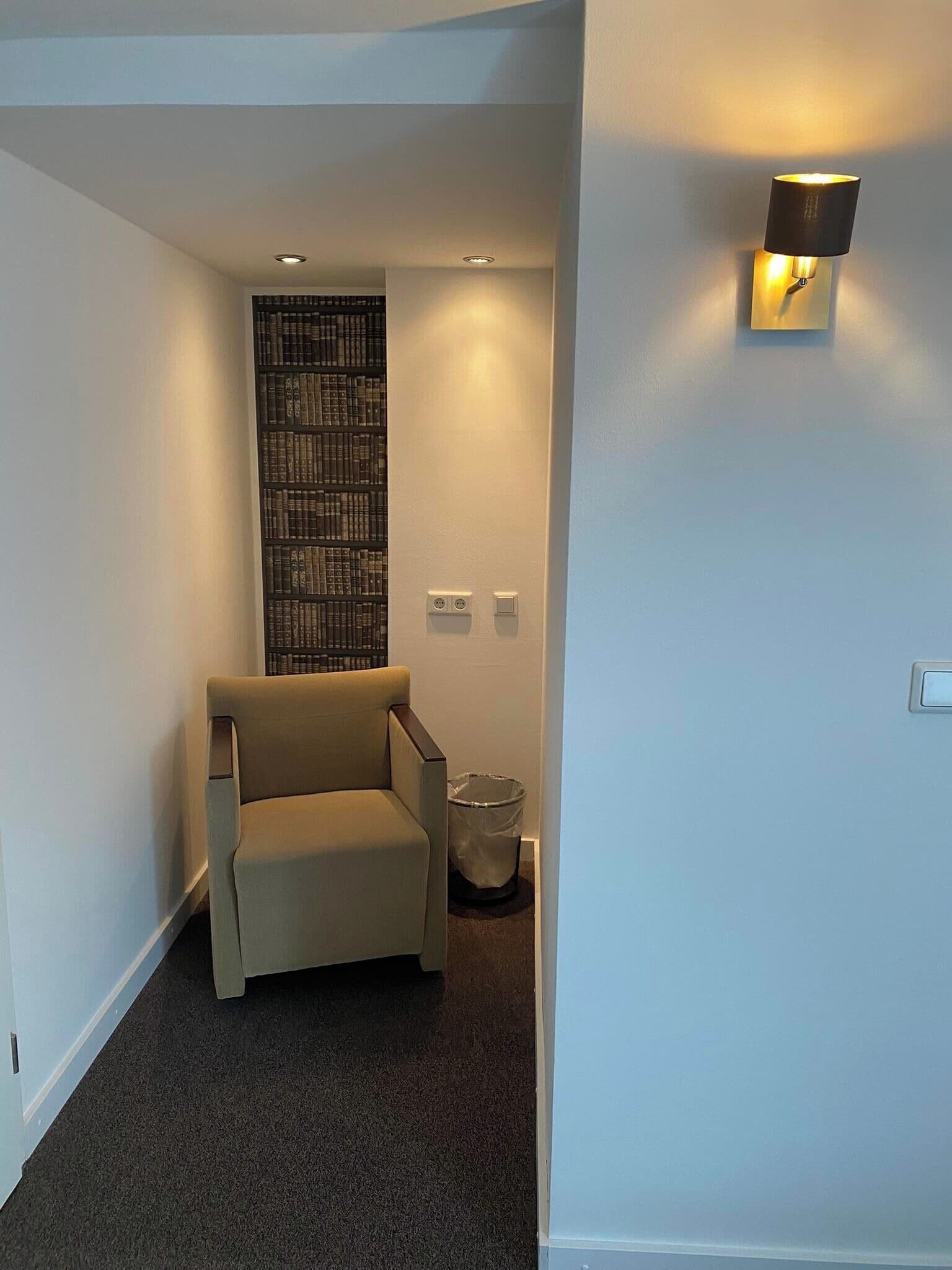 A hallway with a beige armchair, a bookshelf, and a wall-mounted light fixture.