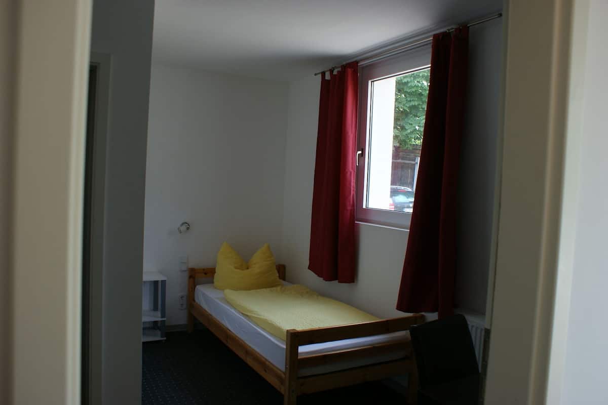 A small, single-bed room with a window, red curtains, and a wooden headboard.