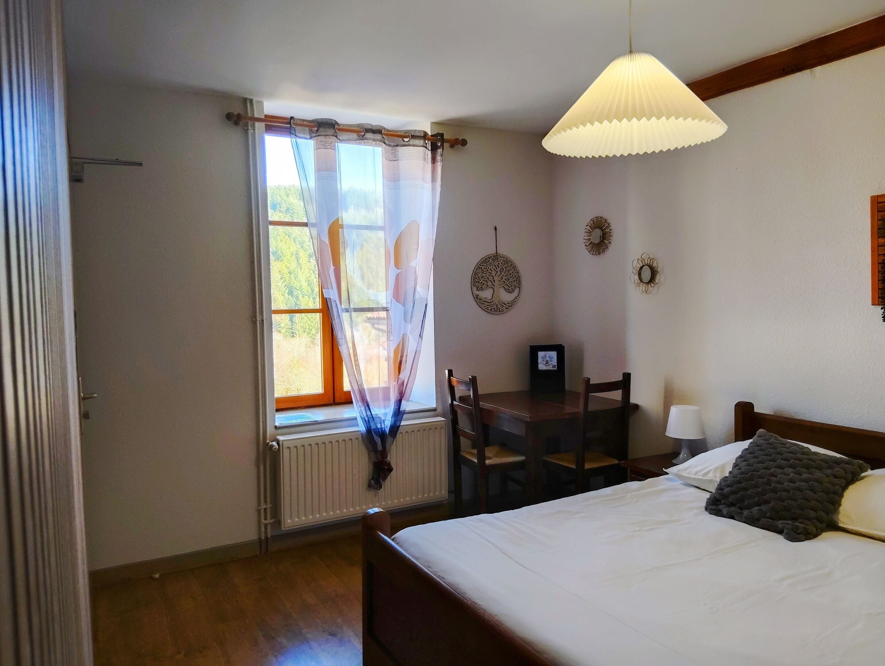 A bedroom with a bed, a desk, and a chair. There is a window with curtains, a radiator, and wall decorations.