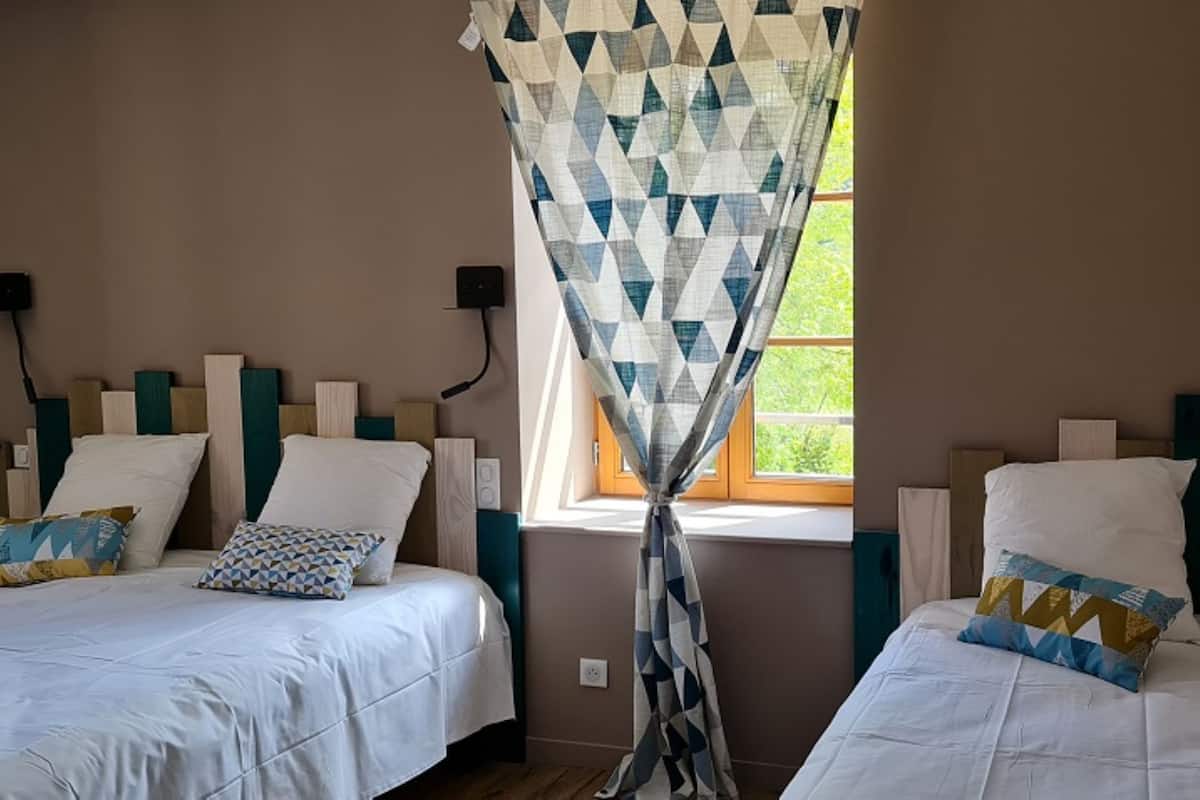 A bedroom with two beds, each with a headboard, a window with a geometric curtain, and wall-mounted lights.