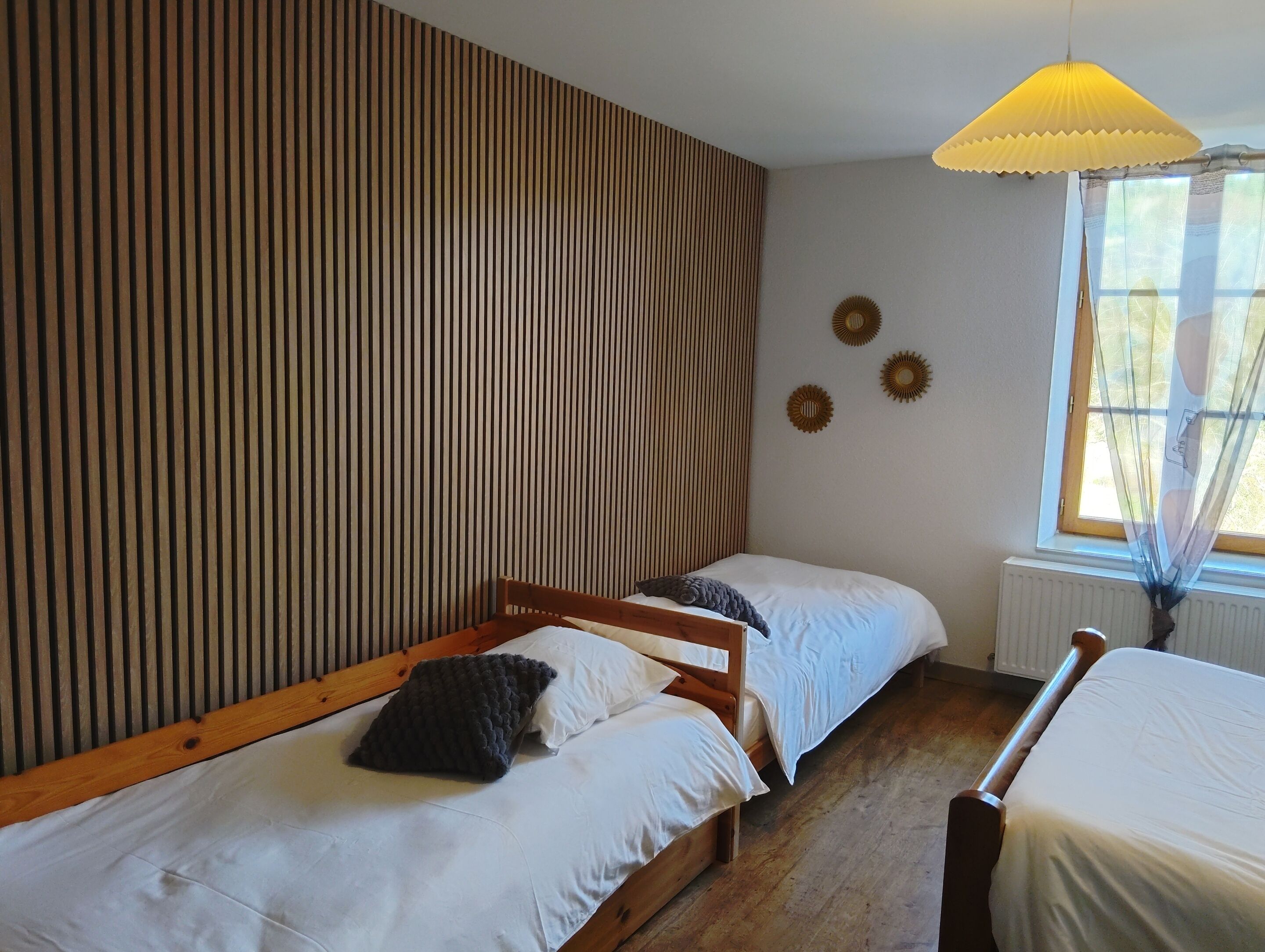 Ein Zimmer mit zwei Holzbettgestellen, einem Fenster mit durchsichtigen Vorhängen und einer Wand mit vertikalen Holzleisten.