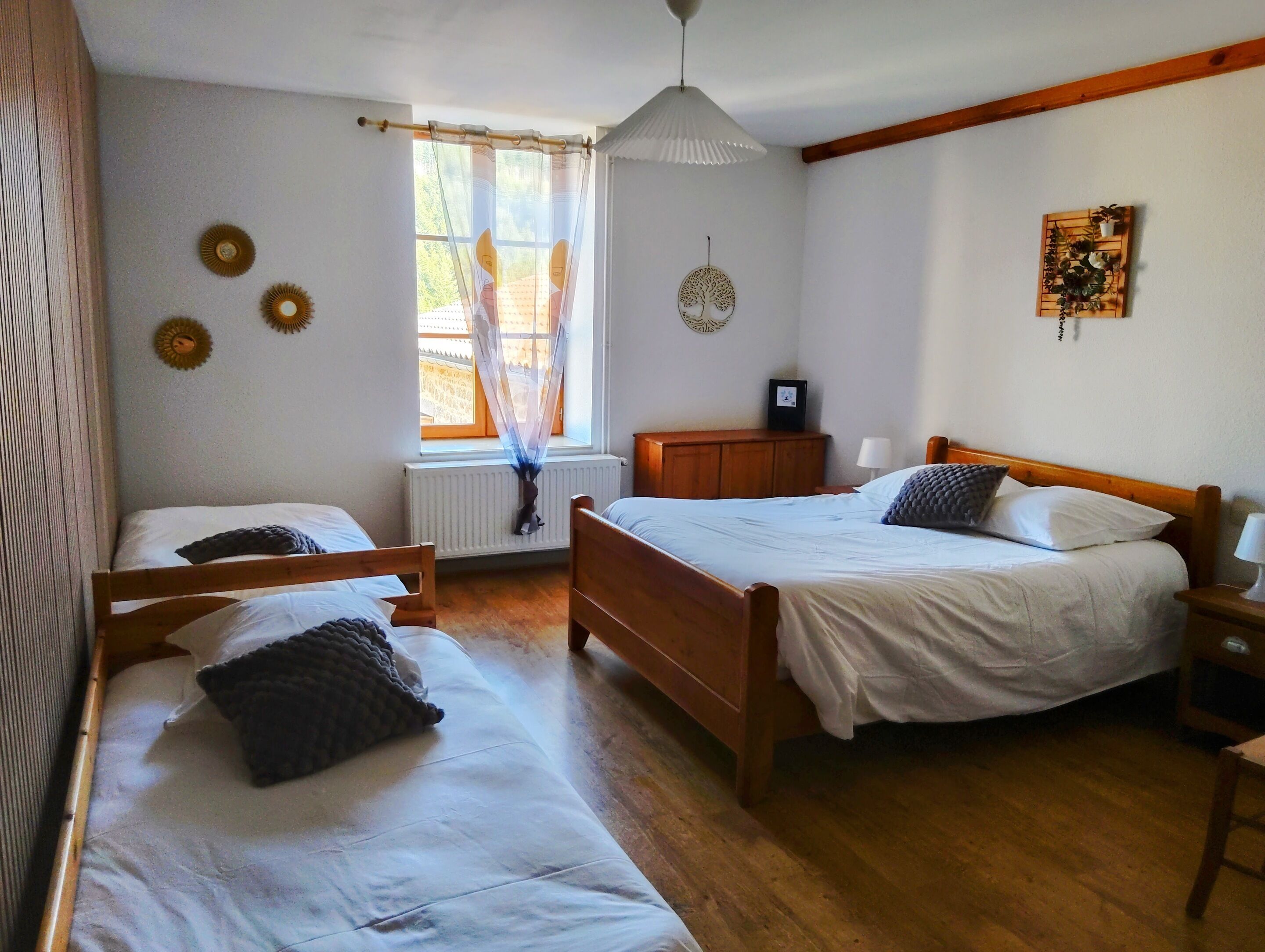 Ein Zimmer mit zwei Betten, einem Fenster mit durchscheinenden Vorhängen, einem Heizkörper, einem Holzbalken an der Decke und einer Wanddekoration.