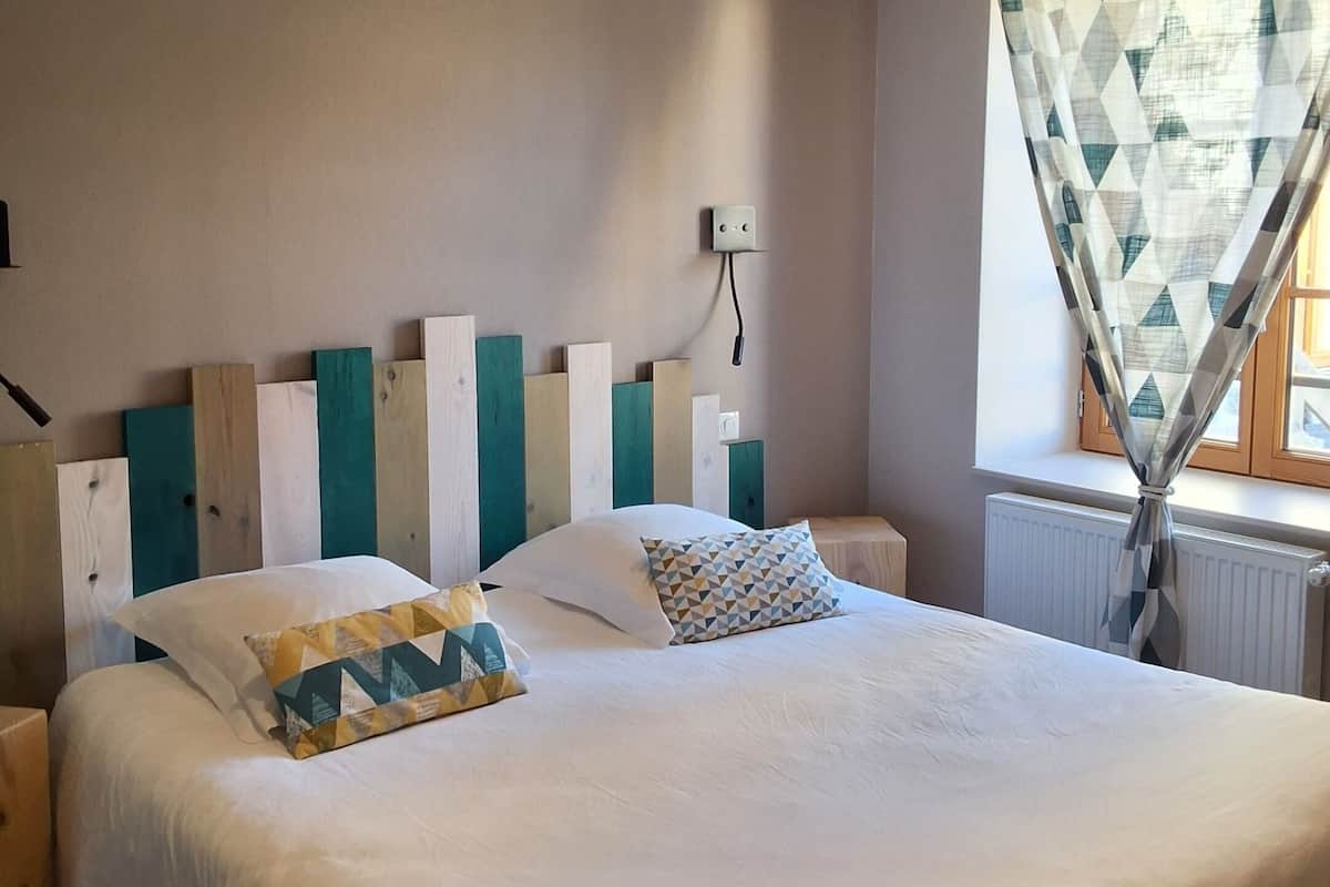 A neatly made bed with white bedding and geometric patterned pillows, a wooden headboard with teal and white panels, a bedside table with a lamp, and a window with geometric patterned curtains.