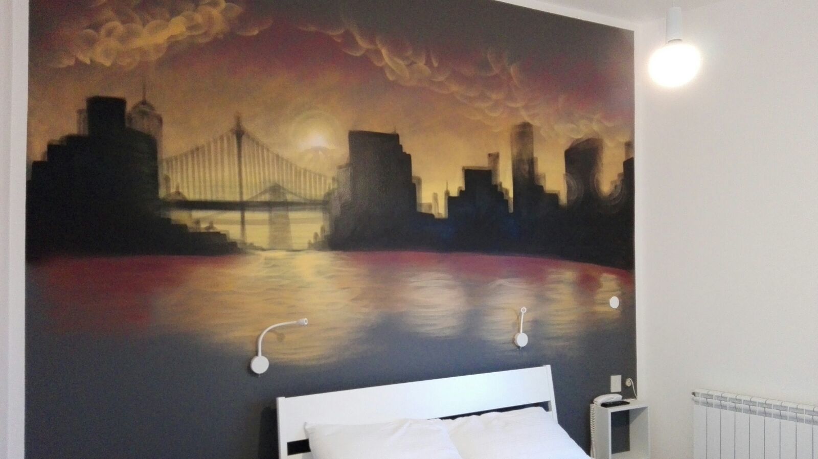 Ein Schlafzimmer mit großem Stadtbild-Wandgemälde, einem weißen Bett und einer Nachttischlampe.