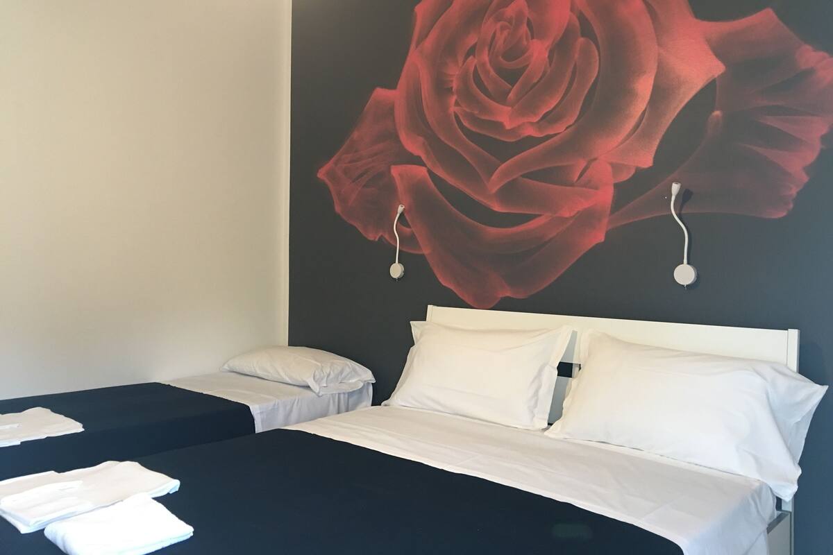 A hotel room with a large wall mural of a red rose, two beds with white linens, and a bedside table with a white lamp.