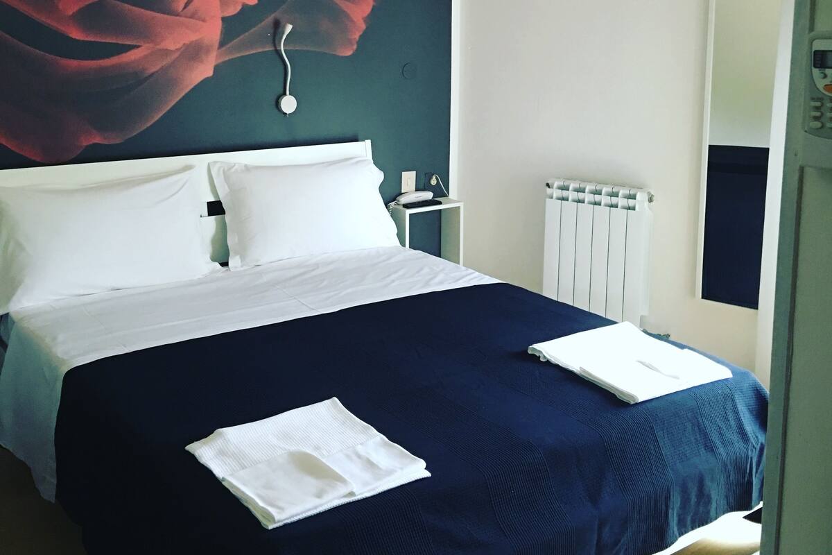 A neatly made bed with white pillows and a dark blue bedspread, a white bedside table with a lamp, and a large red rose mural on the wall.