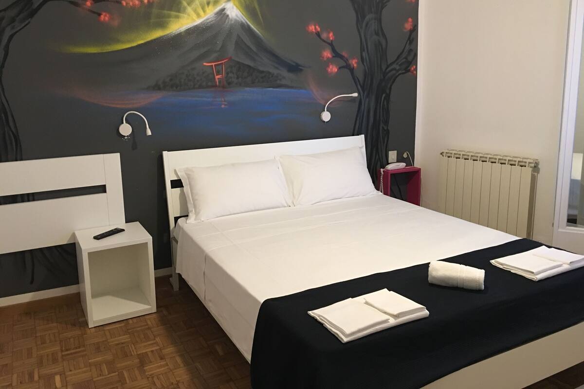 A hotel room with a bed, a nightstand, a red lamp, a white pillow, and a wall mural of a mountain and cherry blossoms.