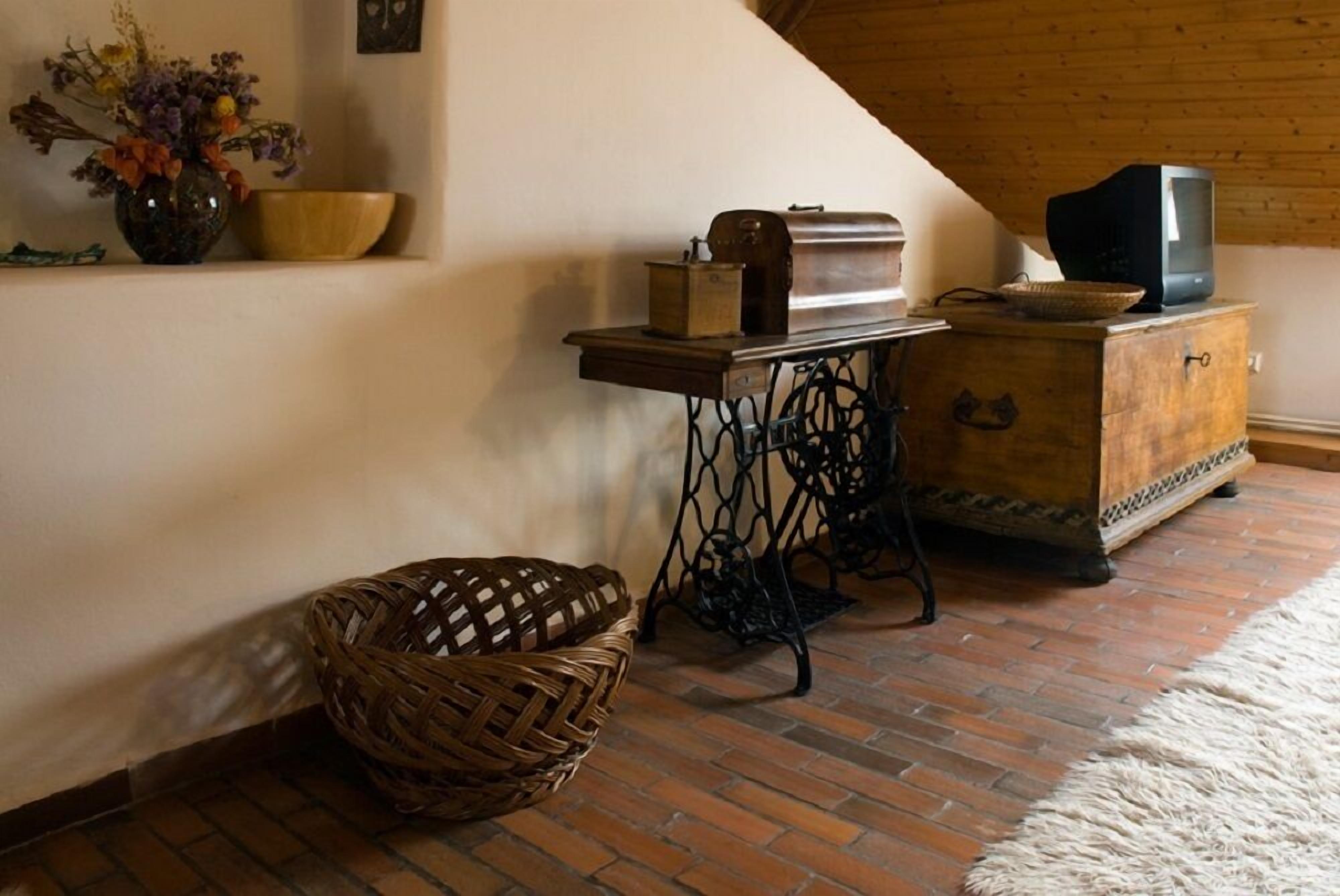 Um quarto com piso de tijolos, uma cesta trançada, uma mesa de madeira com estrutura metálica, um rádio antigo, uma televisão sobre um móvel de madeira e um vaso com flores.