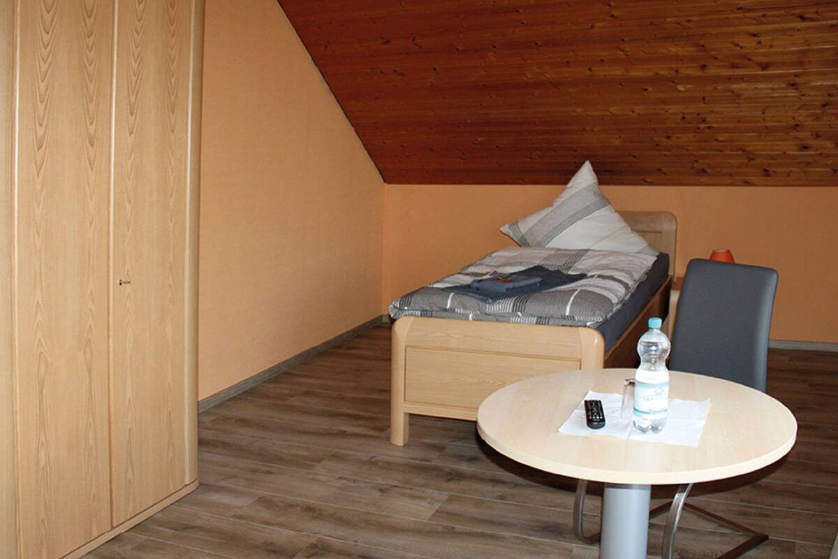 Ein kleiner, gemütlicher Raum mit Holzbalkendecke, einem Bett mit Kissen, einem Stuhl und einem Tisch mit Flasche und Fernbedienung.