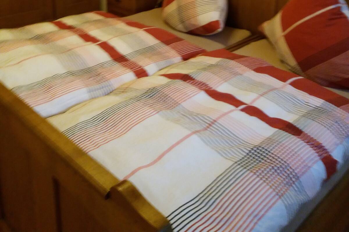 Ein Bett mit kariertem Überwurf und zwei Kissen, ein Holz-Kopfteil, ein Nachttisch mit Lampe und eine Wand mit einem dekorativen Schild.