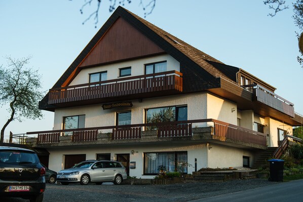 Ein zweistöckiges Gebäude mit einem Balkon im oberen Geschoss, einem Schild mit der Aufschrift 'Bau-Bachwalhof' und einem geparkten Auto davor.