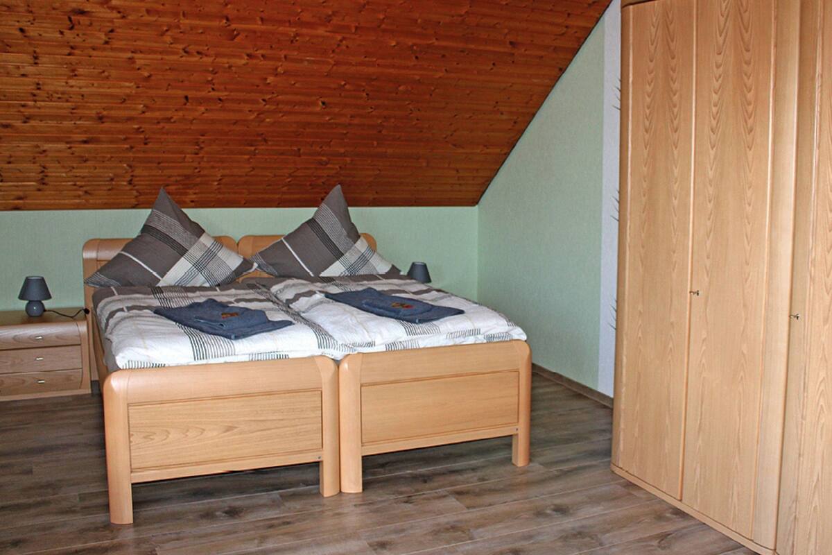 Ein Schlafzimmer mit Holzbalkendecke, einem Bett mit gemustertem Überwurf, zwei Kissen, einem Nachttisch mit Lampe und einem Holzschrank.
