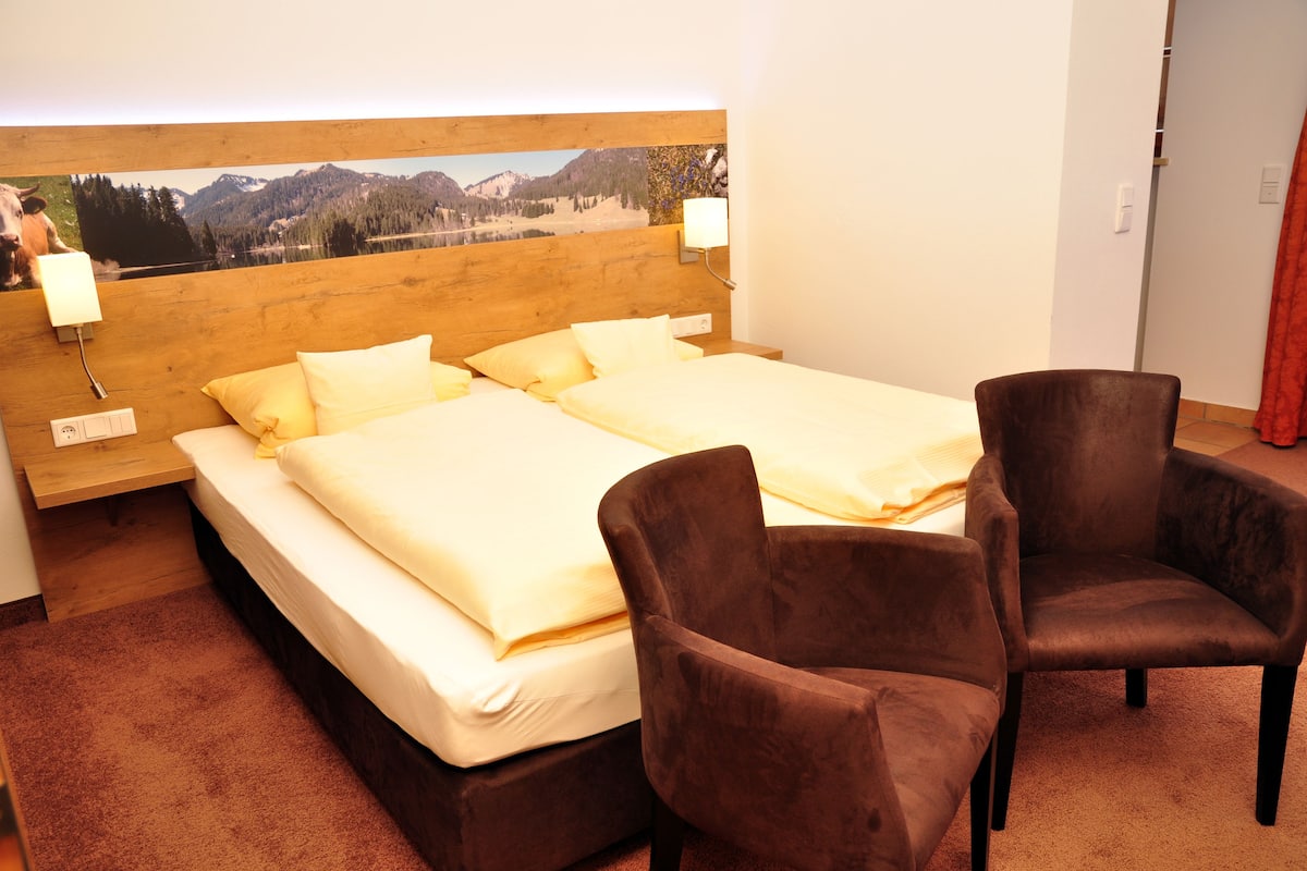 Une chambre d’hôtel avec un grand lit, deux fauteuils marron, une tête de lit en bois et un tableau représentant un paysage de montagne.