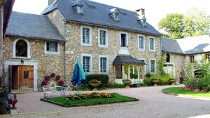 Ein Steinhaus mit einem zentralen Eingang, einer Veranda und einem Garten mit Blumen und einer Statue.