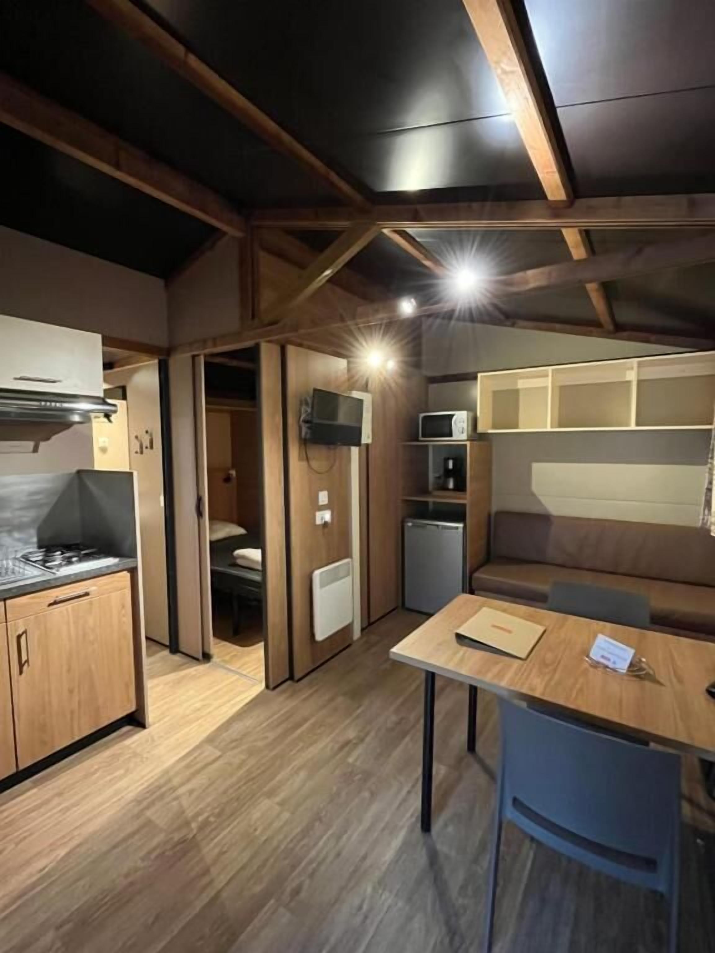 Une cuisine compacte avec des meubles en bois, une cuisinière à gaz, un four à micro-ondes et un petit réfrigérateur. On y trouve une table à manger en bois, une chaise et un banc intégré avec des coussins.