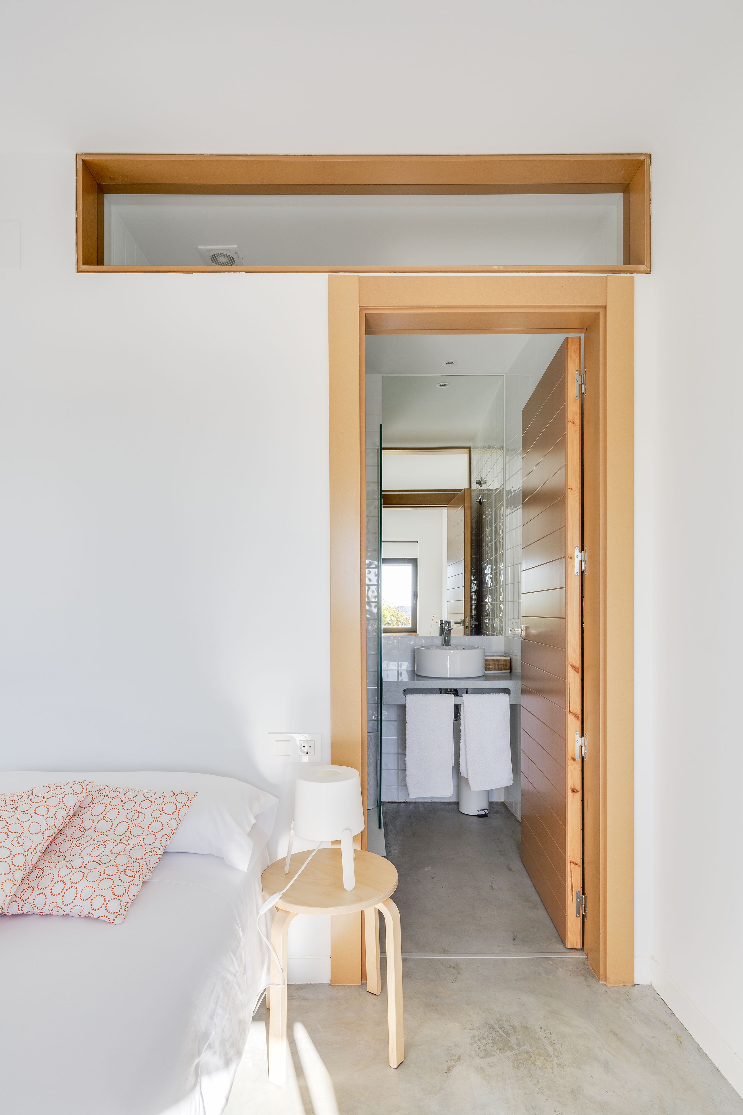 A bedroom with a bed, a bedside table, and a small wooden stool. A bathroom is visible through an open door, featuring a sink, mirror, and towels.
