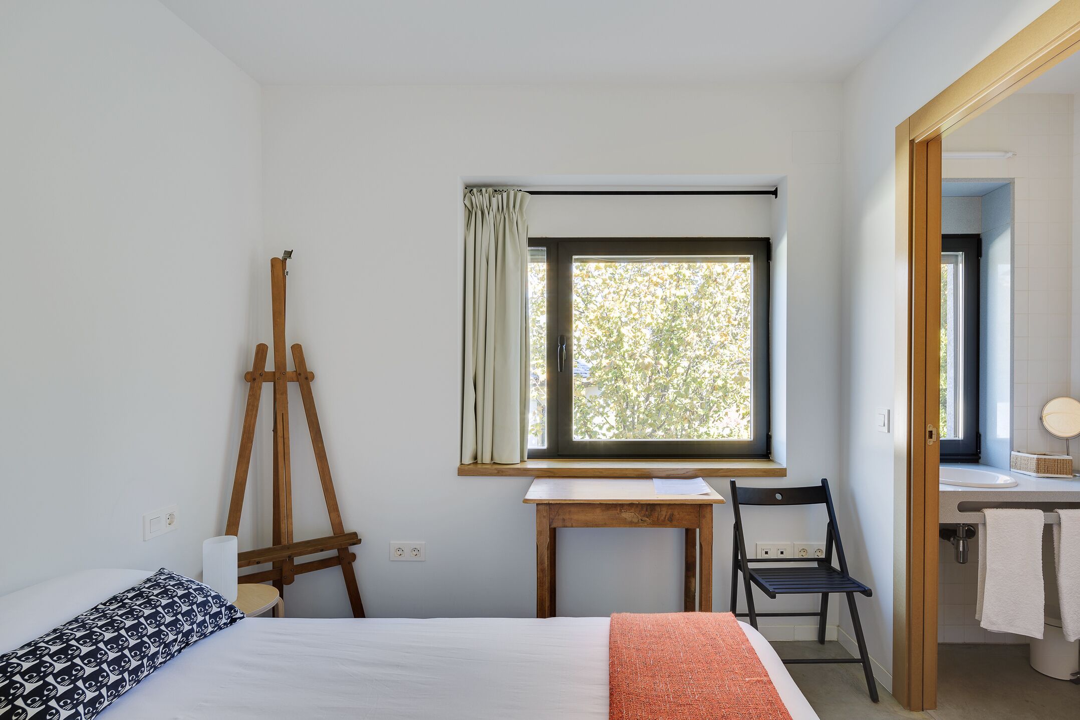 Un dormitorio con una cama, un caballete de madera, una mesita, una silla y una ventana con cortinas.