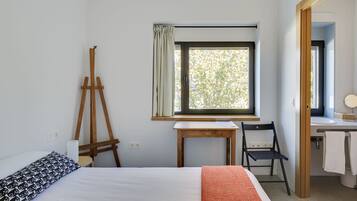 Un dormitorio con una cama, un caballete de madera, una mesita, una silla y una ventana con cortinas.