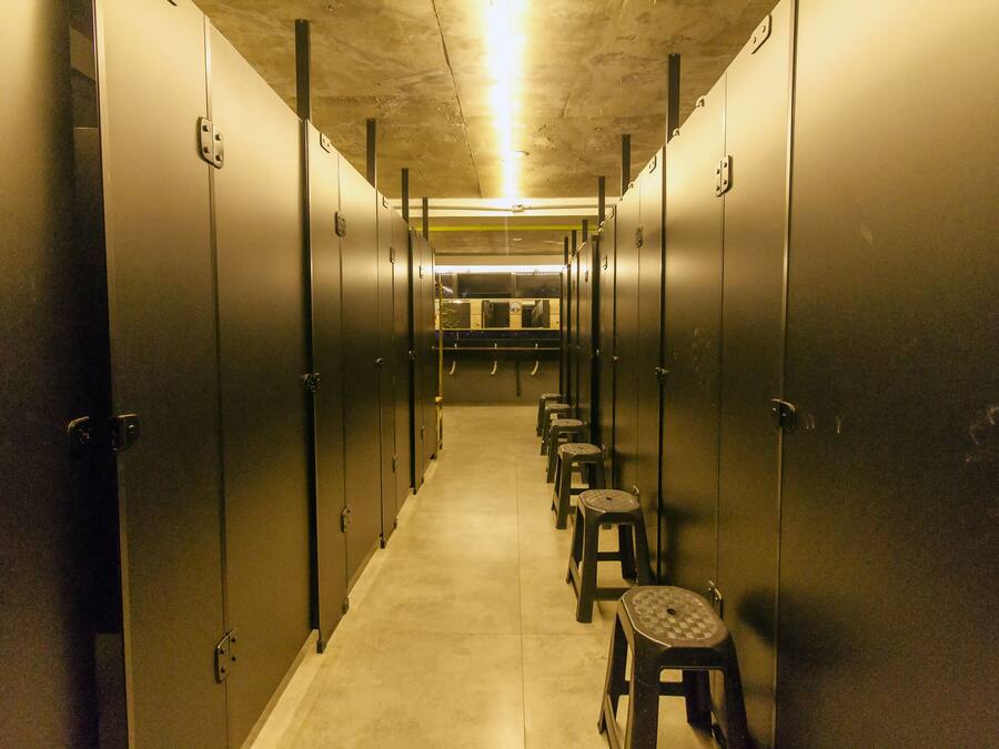 A narrow hallway with metal lockers on both sides and a row of stools in the center.