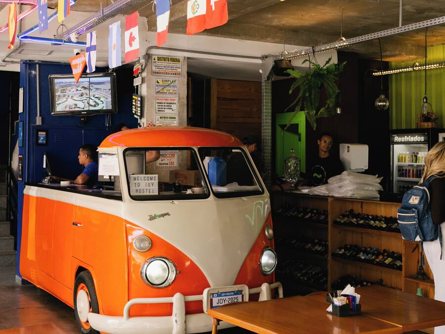 A retro-themed van with a sign reading "WELCOME TO JOY TOSTEL" parked inside a building with a green door and a shoe display.
