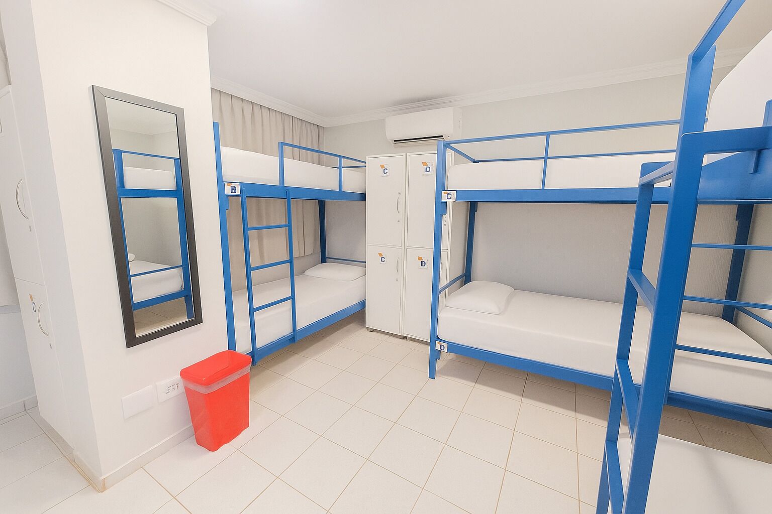 A dormitory room with bunk beds, a mirror, a trash bin, and a wardrobe.