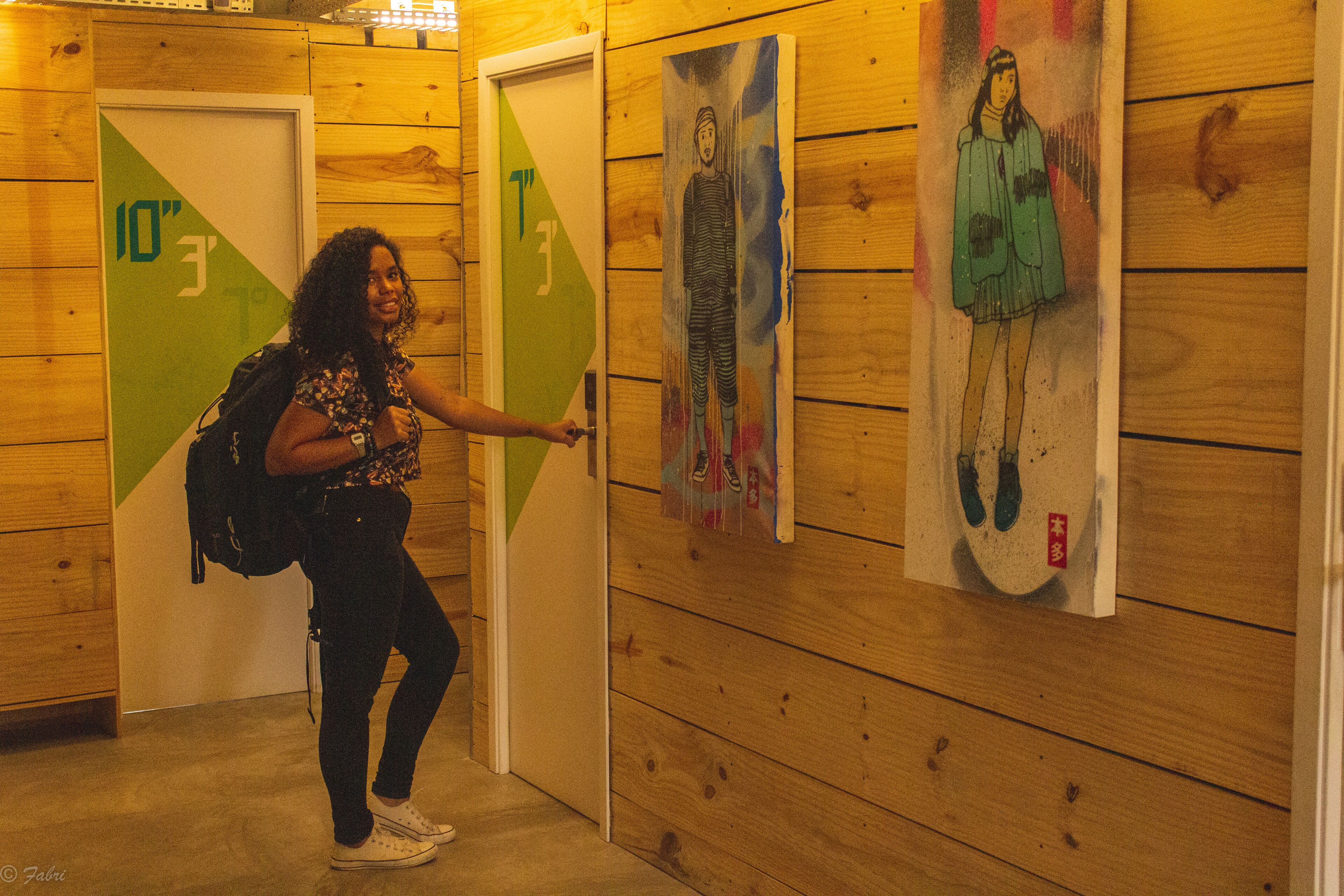 Una persona de pie en un pasillo con paredes y puertas de madera, señalando una pared con obras de arte coloridas.