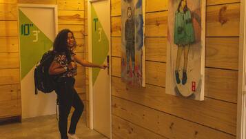 Una persona de pie en un pasillo con paredes y puertas de madera, señalando una pared con obras de arte coloridas.