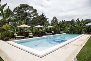 A rectangular swimming pool with a tiled edge, surrounded by a wooden deck. There are lounge chairs and umbrellas along the poolside.