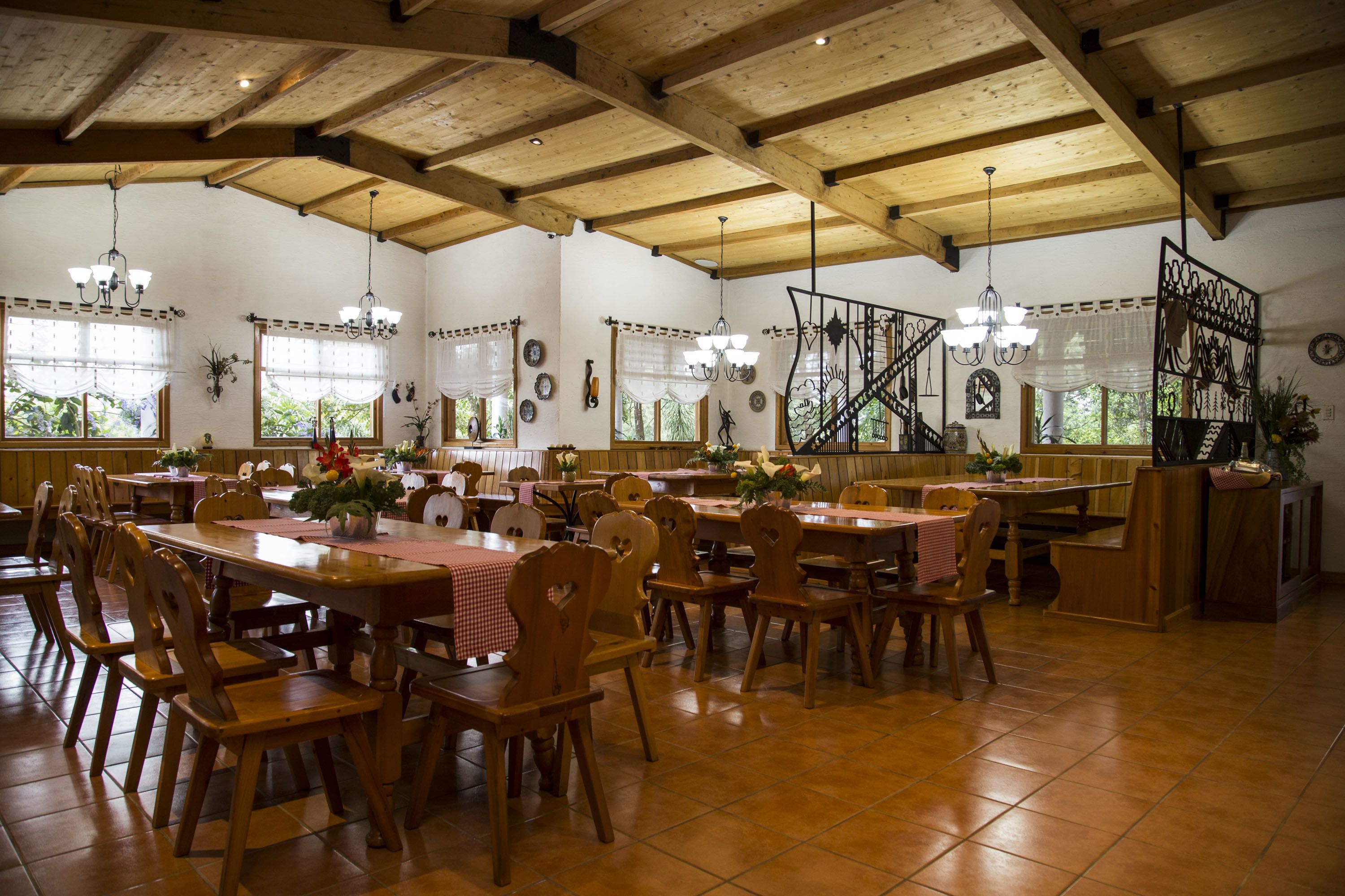 A spacious dining area with wooden tables and chairs, large windows with curtains, and a high wooden ceiling.