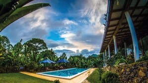 A swimming pool with a stone wall and a covered area with blue umbrellas. - Hotel Boutique Y Restaurante Panisté (San Cristobal Verapaz)
