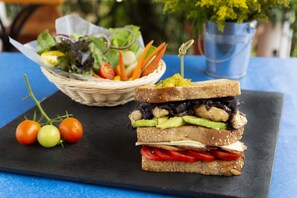 A sandwich with various ingredients on a black slate board, a basket of salad, and some cherry tomatoes on the side.