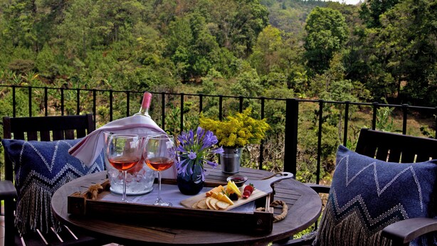 A table with a bottle of wine, glasses, a cheese board, and a flower arrangement on a balcony with a view of a forested hillside. - Hotel Boutique Y Restaurante Panisté (San Cristobal Verapaz)
