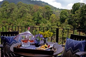 Una mesa con una botella de vino, copas, una tabla de quesos y un arreglo floral en un balcón con vistas a una ladera boscosa.