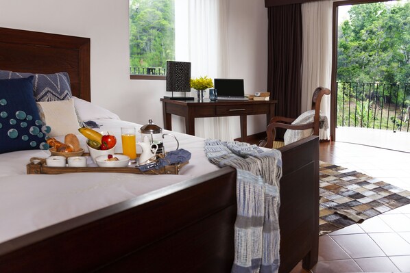 A neatly made bed with breakfast items, a laptop on a desk, and a view of greenery through a balcony.