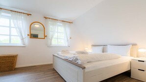 A bedroom with a white bed, a wooden headboard, a nightstand with a lamp, and a wicker basket. - Holiday house Witte Hus (ID 175) - Holiday house "Witte Hus" (ID 175) (Sankt Peter-Ording)