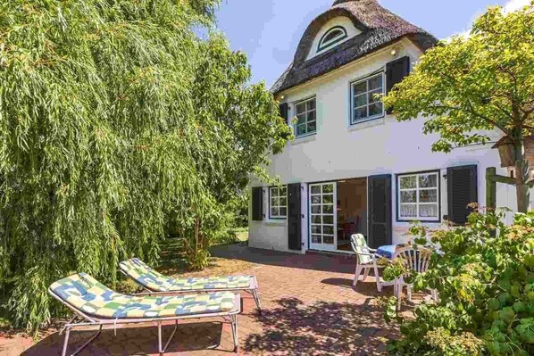 Une maison à deux étages avec un toit de chaume, un grand arbre au premier plan et deux chaises longues dans le jardin.
