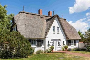 Une maison blanche avec un toit de chaume à forte pente et un petit porche agrémenté de plantes en pot.