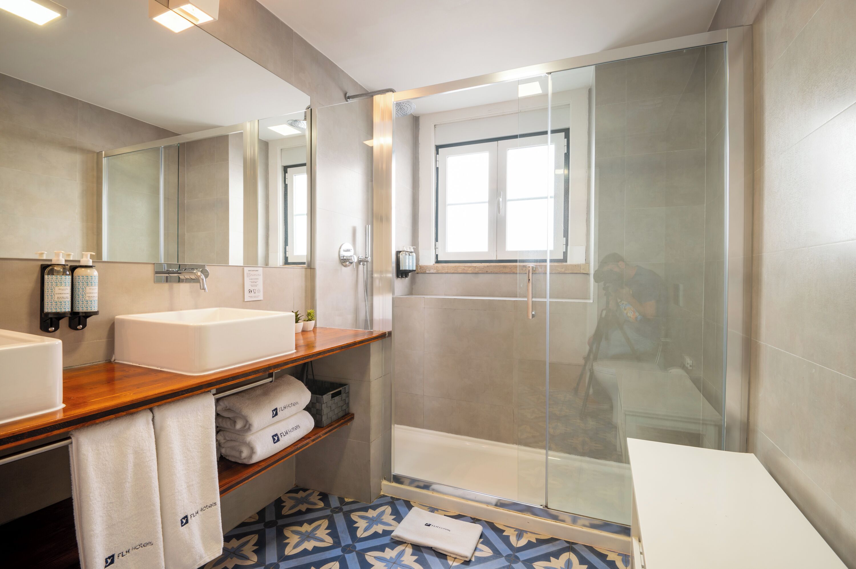A modern bathroom with a wooden vanity, two sinks, a glass shower enclosure, and a patterned floor.