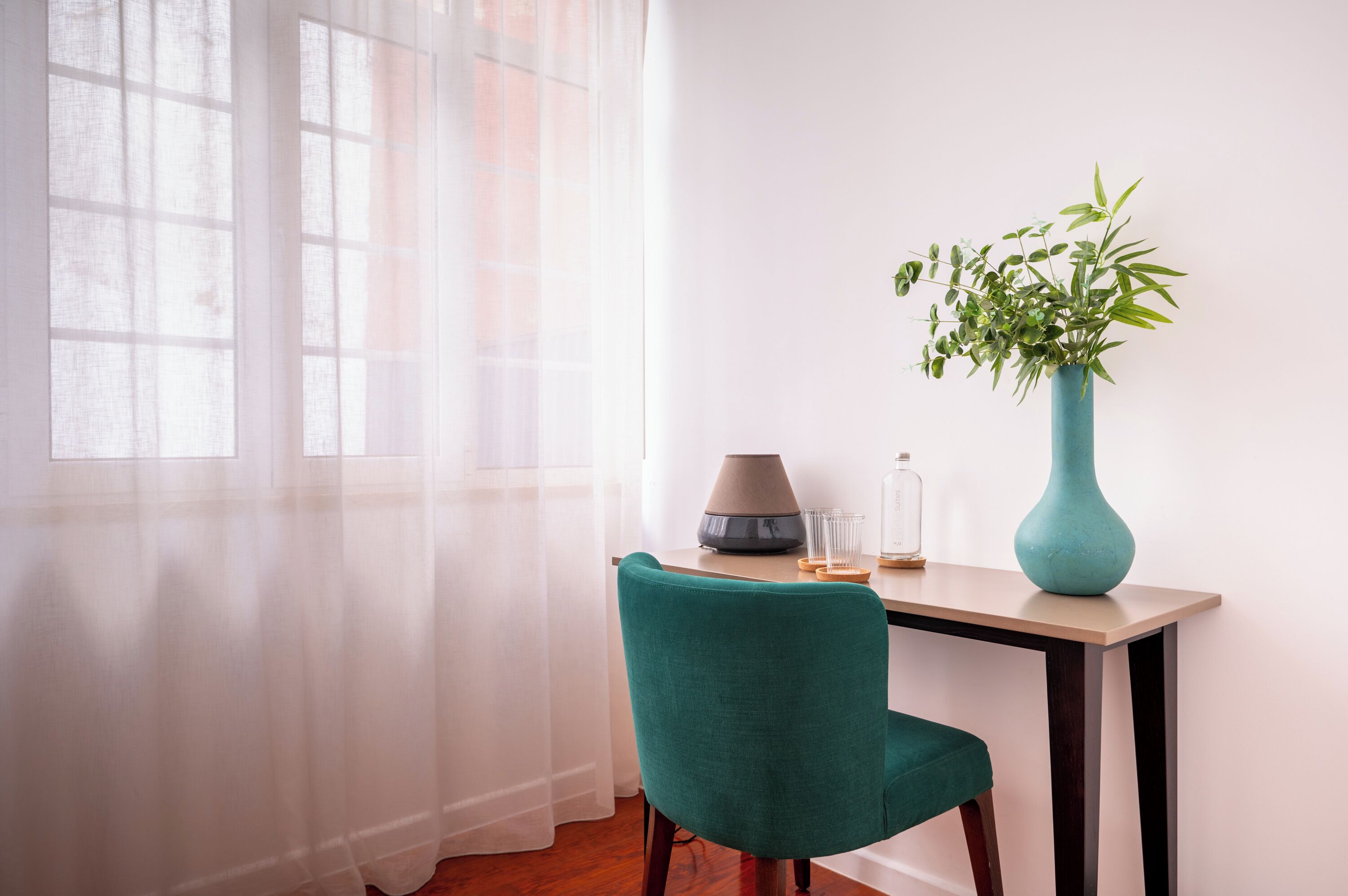 A room with a desk, a chair, a lamp, a vase with a plant, and sheer curtains.