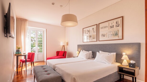 A hotel room with two beds, a desk with a chair, a bench, a TV, and framed pictures on the wall. - Bica FLH Suites (Lisbon)