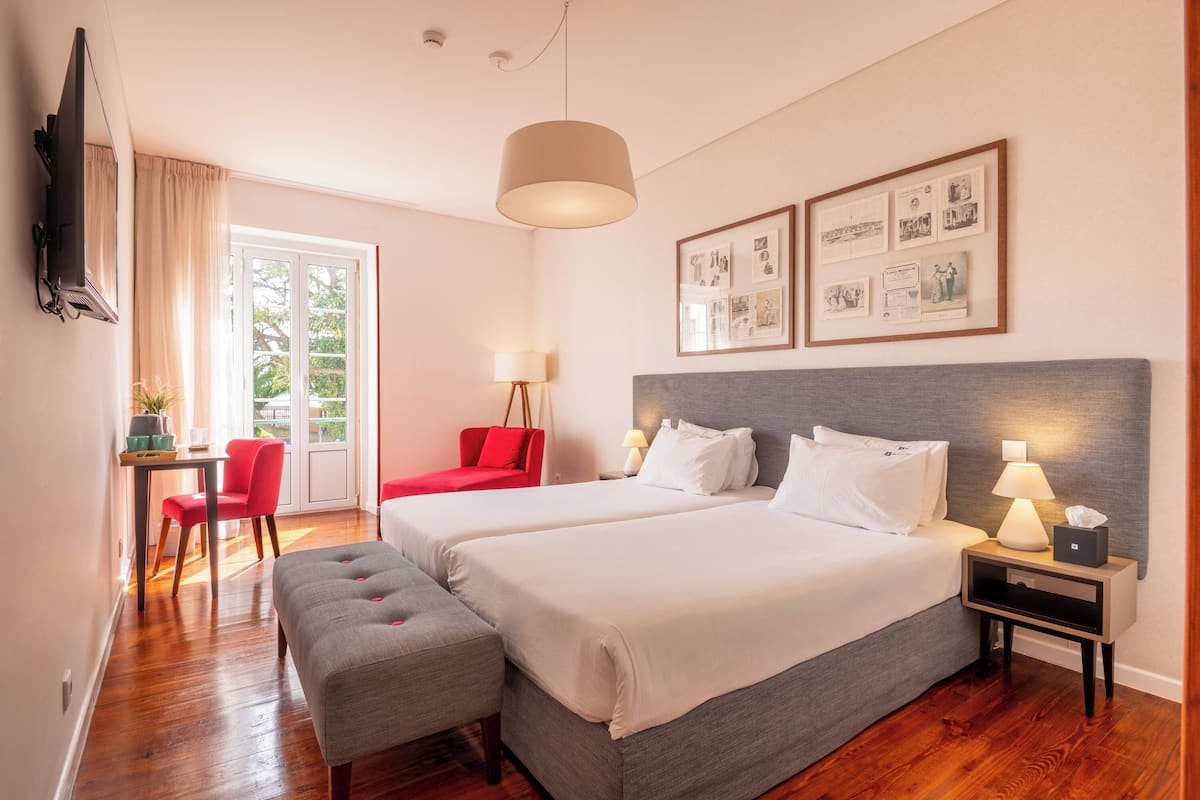 Habitación de hotel con dos camas, un escritorio con silla, un banco, televisor y cuadros en la pared.