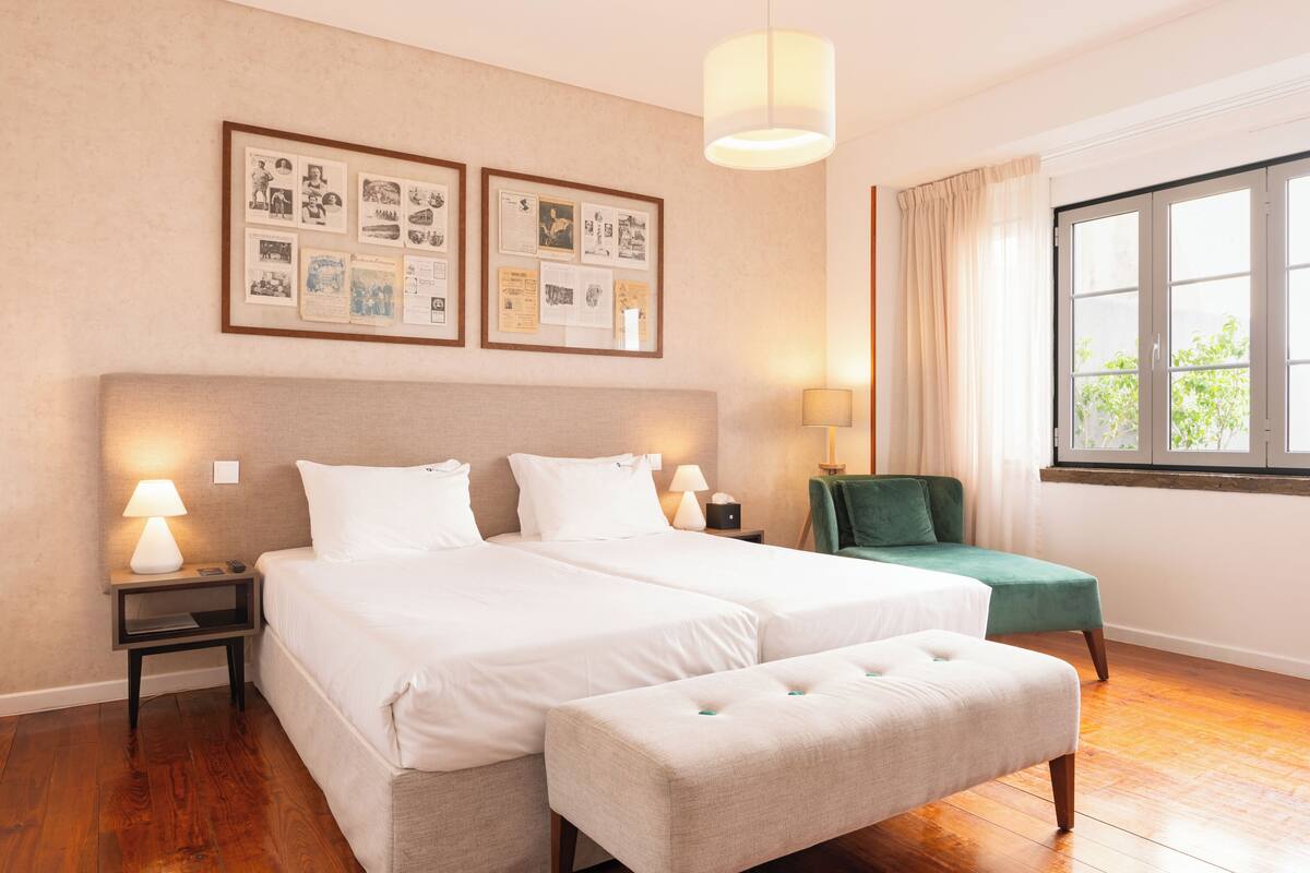 Una cama doble con dos almohadas, una mesita de noche con lámpara, una silla verde y una ventana con cortinas.