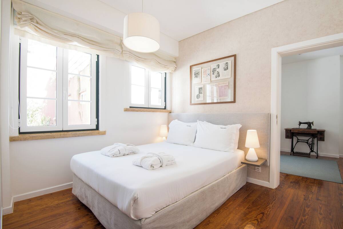 Una cama bien hecha con sábanas blancas y dos toallas, una mesita de noche con una lámpara, una ventana con cortinas y un cuadro en la pared.